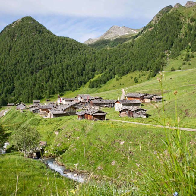 Find your hotel near Brixen! The picture shows an idyllic mountain village with wooden houses, located in a green valley. In the foreground flows a small brook, crossed by a wooden bridge, and in the background wooded mountains are visible.