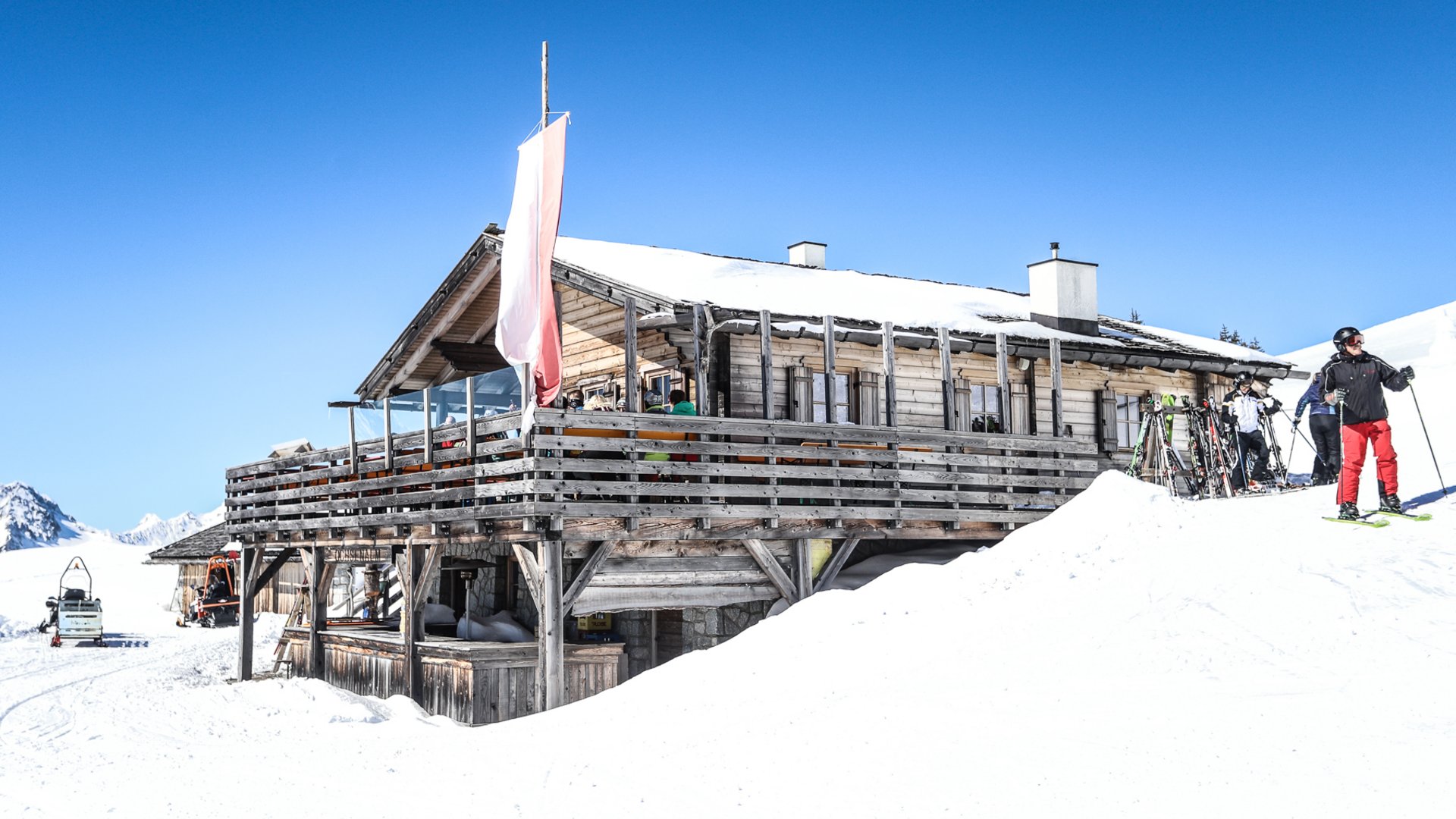 Gitschberg Jochtal holiday area – mountain huts and shelters The picture shows a rustic wooden ski hut surrounded by snow, with some guests sitting on its terrace; in the foreground, a skier stands next to skis that have been put down.