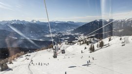 Skigebied Gitschberg Jochtal De foto toont een uitgestrekt skipiste met tal van skiërs die de helling afrijden. Boven de piste loopt een kabelbaan met gondels, en op de achtergrond strekt zich een indrukwekkend panorama uit van met sneeuw bedekte bergen en valleien onder een heldere blauwe lucht.