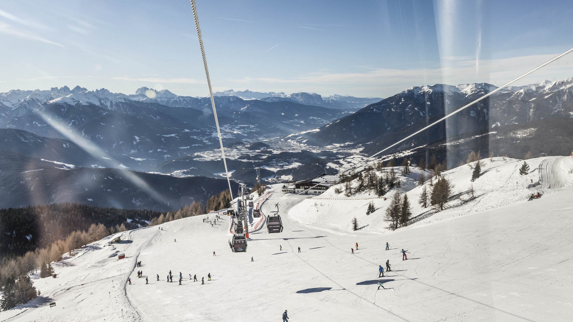 Skigebied Gitschberg Jochtal De foto toont een uitgestrekt skipiste met tal van skiërs die de helling afrijden. Boven de piste loopt een kabelbaan met gondels, en op de achtergrond strekt zich een indrukwekkend panorama uit van met sneeuw bedekte bergen en valleien onder een heldere blauwe lucht.