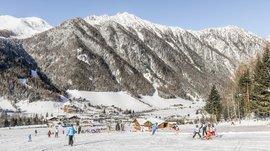 Vals: das Almendorf Das Bild zeigt eine verschneite Berglandschaft mit steilen Berghängen im Hintergrund und Skifahrern im Vordergrund. Mehrere Menschen fahren den Hang hinunter, während sich im Tal ein kleines Dorf mit Holzhäusern erstreckt.
