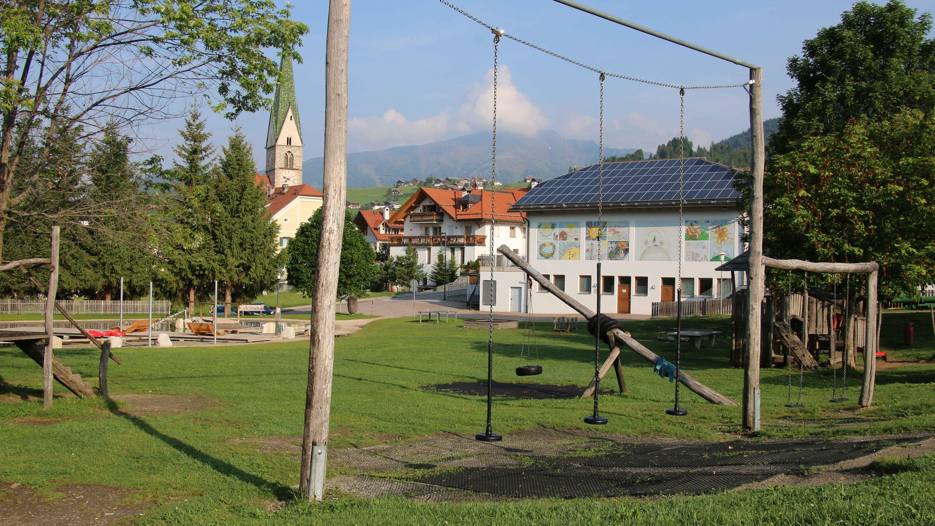 Aktivpark Terenten Obrázek zobrazuje hřiště s houpačkami v popředí, které leží na zelené louce. V pozadí je budova s solárními panely, malovanými stěnami a kostel s ostrou věží, obklopený stromy a kopcovitou krajinou.