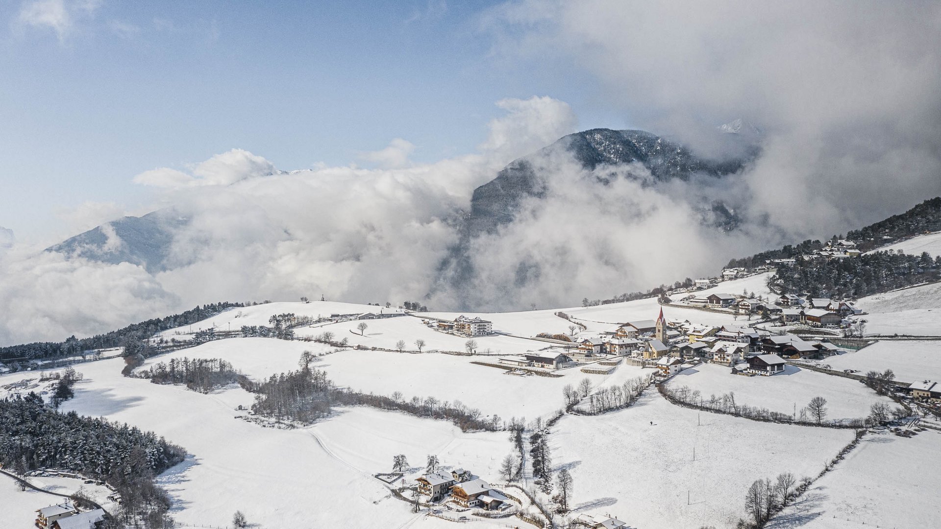 The Alpine hamlet of Spinges The picture shows a snowy village located in a gently rolling landscape. The mountains in the background are partly shrouded in clouds, and the sky is clear blue, making the winter landscape appear bright and peaceful.