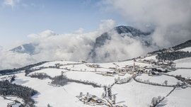 The Alpine hamlet of Spinges The picture shows a snowy village located in a gently rolling landscape. The mountains in the background are partly shrouded in clouds, and the sky is clear blue, making the winter landscape appear bright and peaceful.