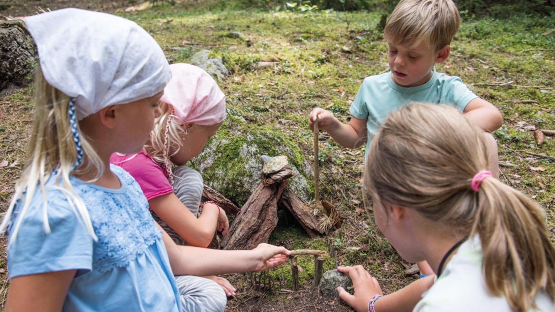 Elfí cesta (Elfenweg) u Vintlu Obrázek ukazuje čtyři děti, které si spolu hrají v lese. Staví malou stavbu ze špejlí, kamenů a kůry na zemi, obklopenou mechem a stromy.