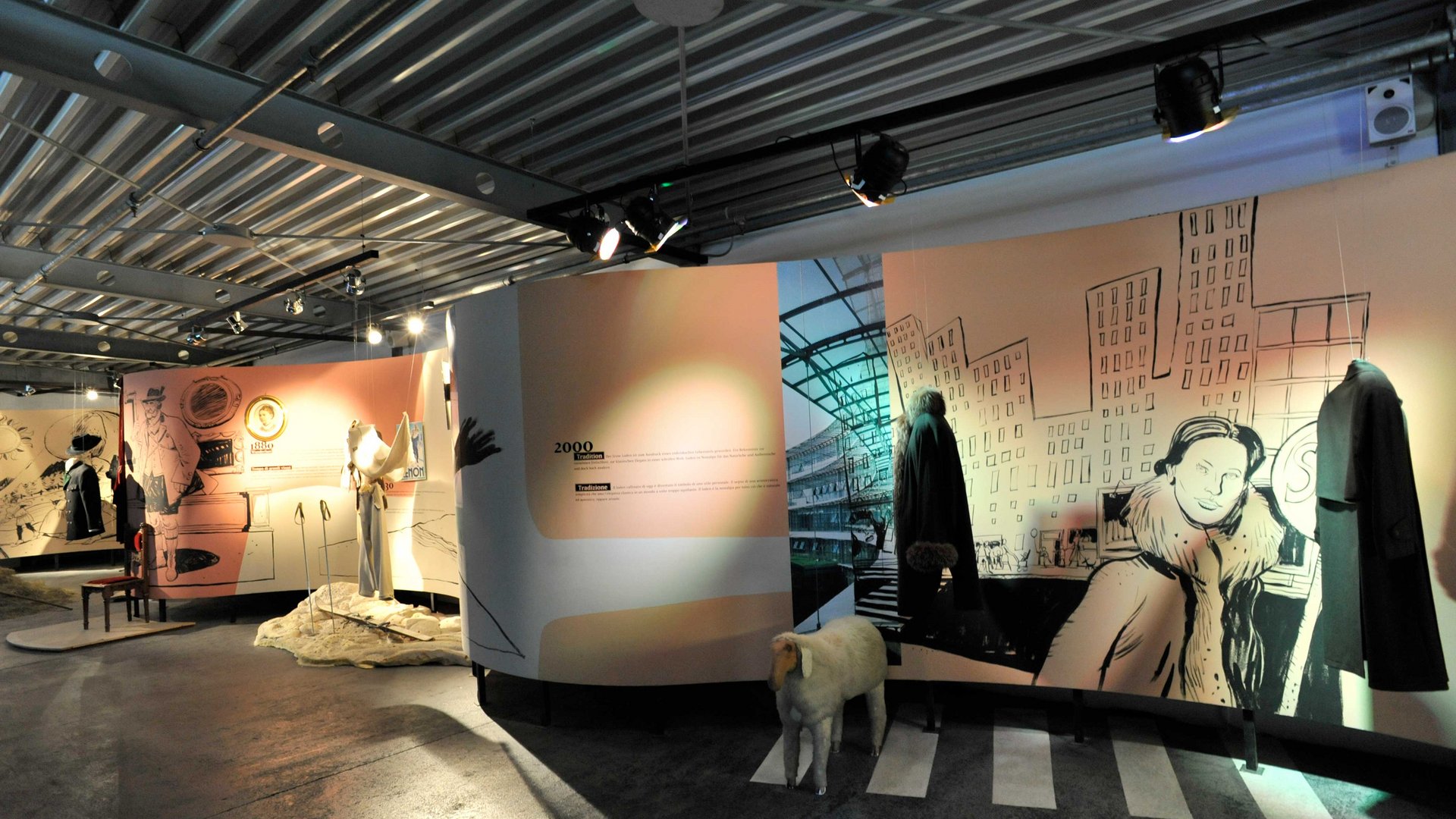 Zážitkový svět lodenu ve Vintlu/Pustertal Obrázek zobrazuje muzeální instalaci, ve které jsou vystaveny historické oděvy. Na stěnách jsou ilustrace scén z různých epoch, včetně městského pohledu. Před stěnami stojí figuríny oblečené v kabátech a dalších oděvech, a na podlaze je model ovce.