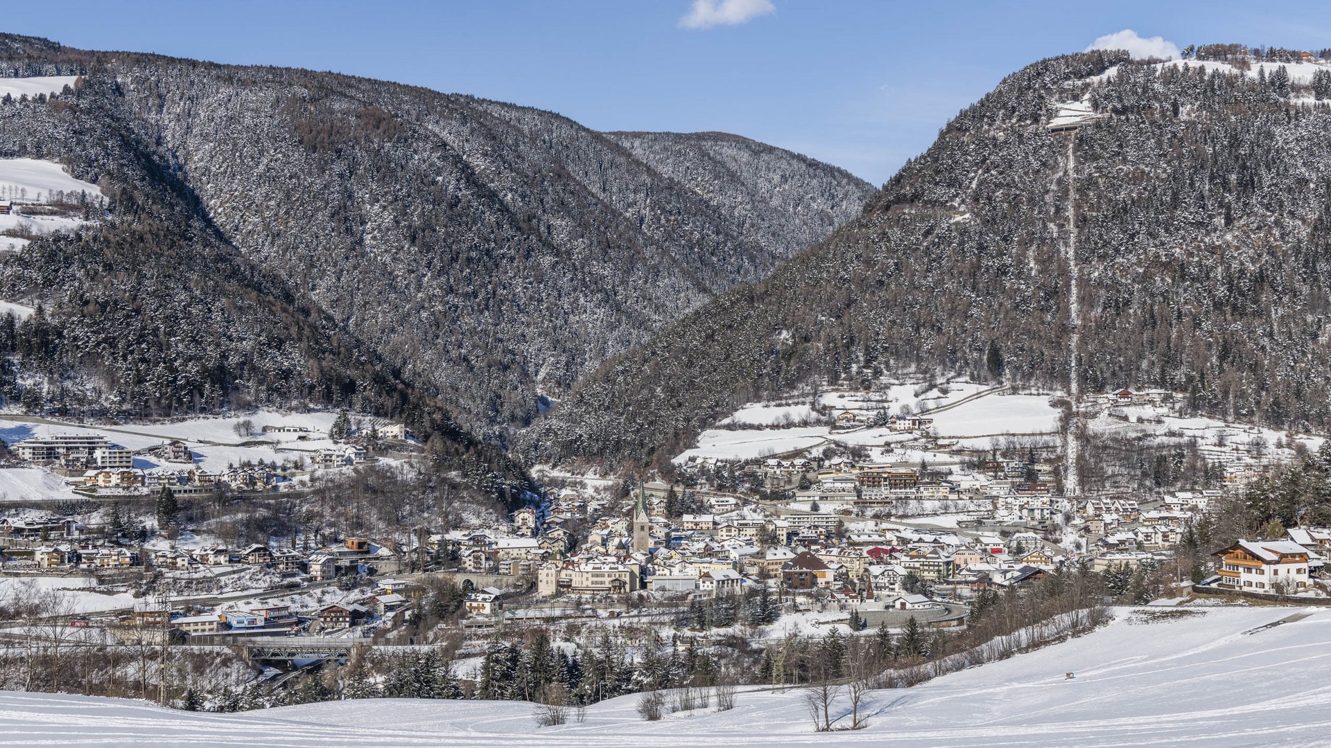 Mühlbach in the Pustertal valley The picture shows a city located in a snow-covered valley, surrounded by densely forested hills. The buildings are covered with snow, and in the background, snowy hills and a clear blue sky can be seen.