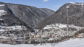 Mühlbach in the Pustertal valley The picture shows a city located in a snow-covered valley, surrounded by densely forested hills. The buildings are covered with snow, and in the background, snowy hills and a clear blue sky can be seen.