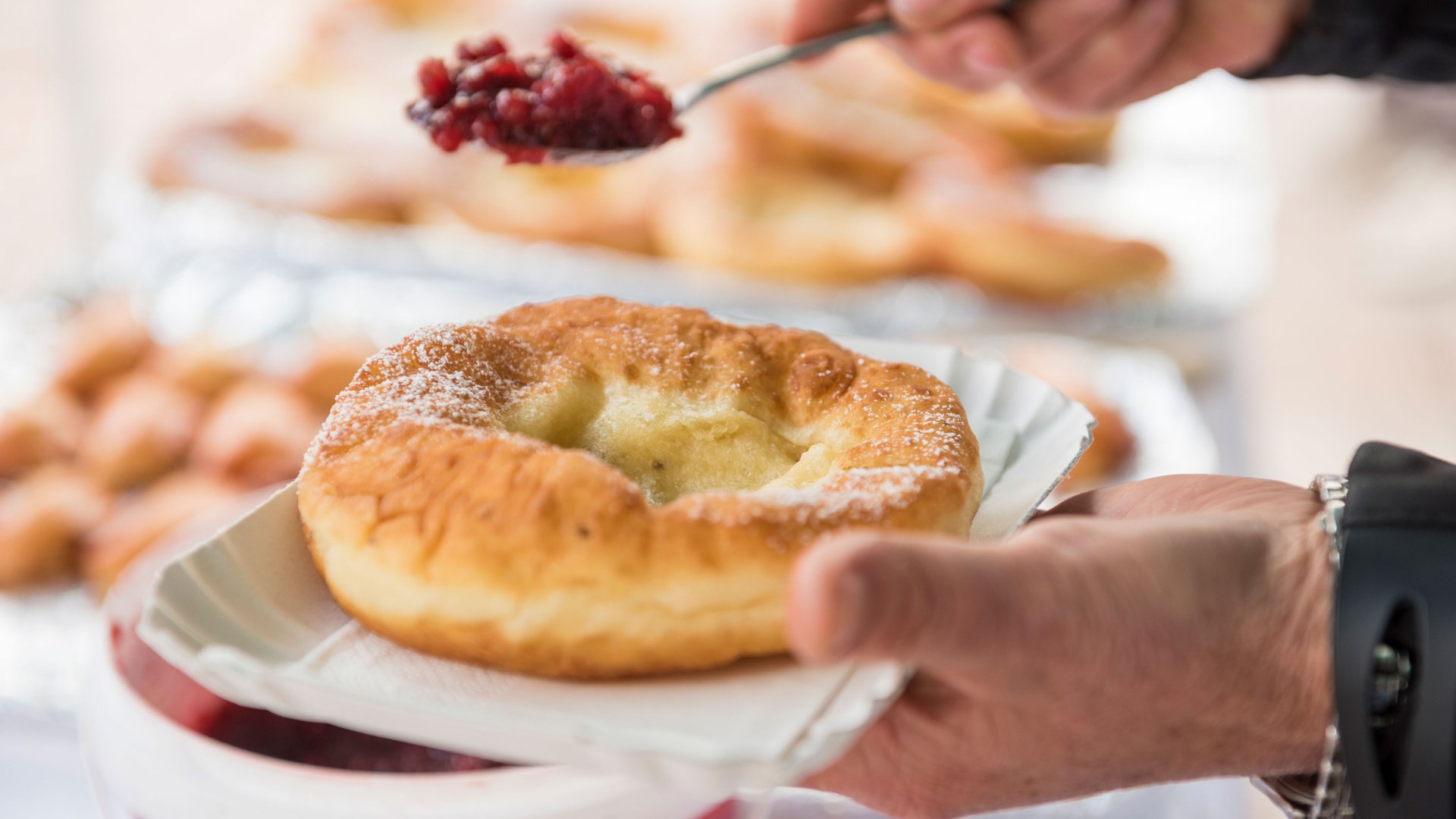 Nakupování v Mühlbachu/Itálie Obrázek ukazuje osobu, která právě lžičkou nanáší červený džem nebo kompot na smažené pečivo, které drží v ruce na papírovém talířku. Pečivo je posypané moučkovým cukrem a vypadá zlatavě hnědé a křupavé.