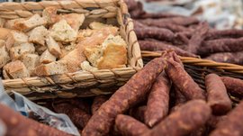 Nakupování v Mühlbachu/Itálie Obrázek zobrazuje koš s kousky chleba, které jsou rustikálně nakrájené. Vedle koše leží mnoho kořeněných, vzduchem sušených klobás v další pletené bedýnce. Scéna naznačuje tradiční tržní nebo farmářské prostředí.