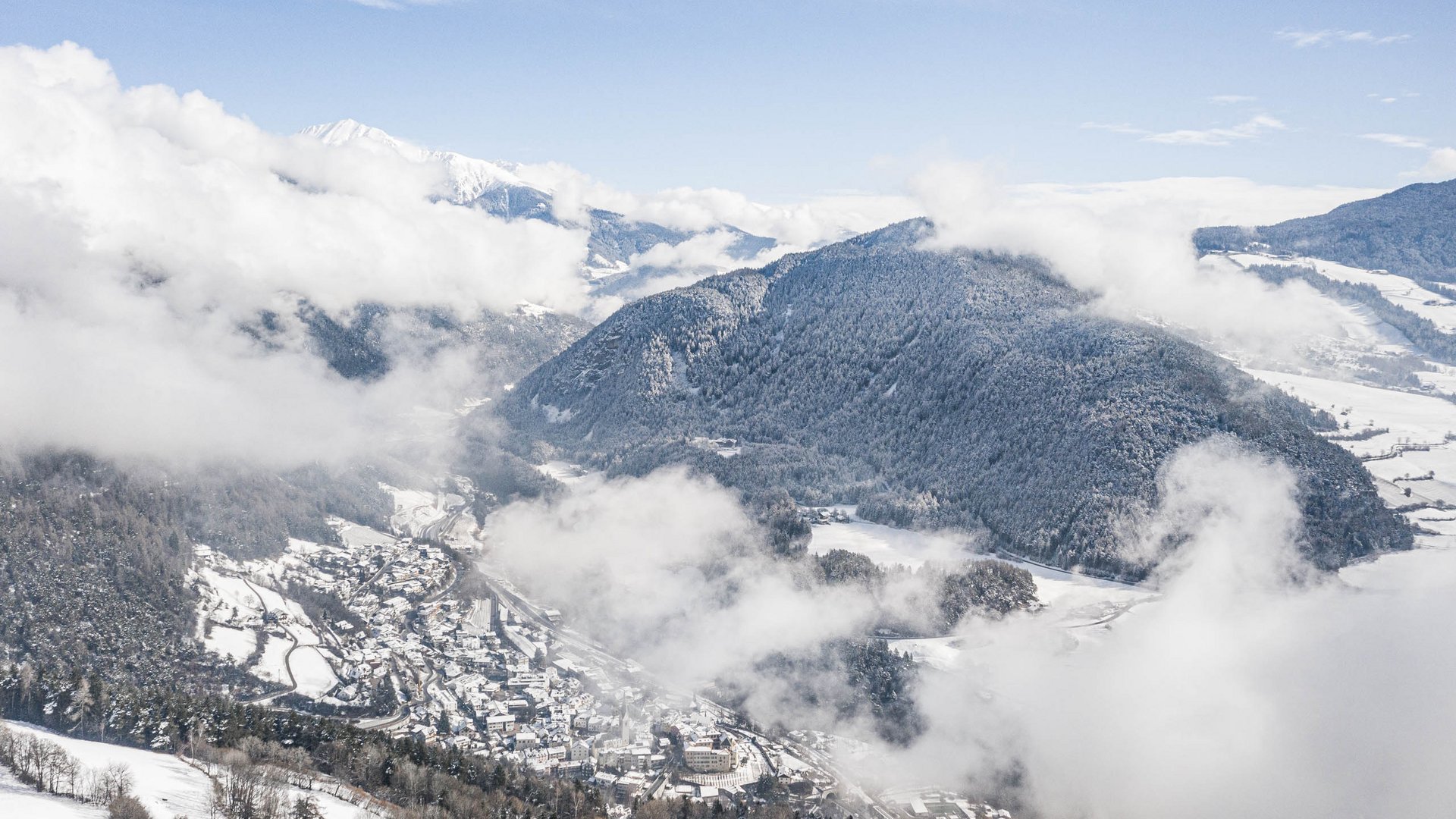 Mühlbach in the Pustertal valley The picture shows a snowy landscape with a valley and a small town surrounded by wooded hills and mountains. Clouds drift over the scene, obscuring parts of the landscape and creating a picturesque, wintry impression.