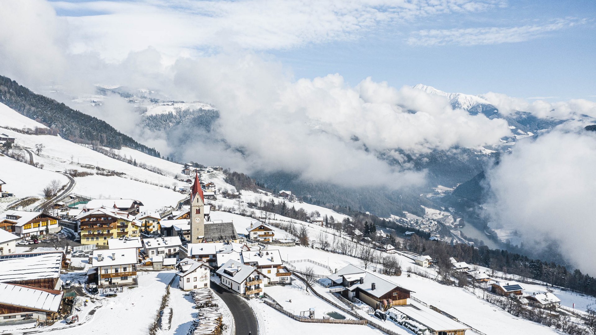 The Alpine hamlet of Spinges The picture shows a snowy mountain village with a church whose red tower stands out among the snow-covered houses. In the background, you can see wooded hills and low-hanging clouds that partially obscure the valley below.