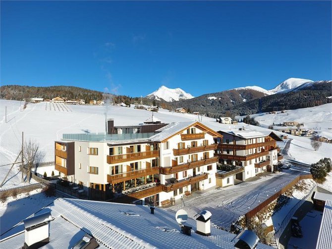 Willkommen in der Ferienregion Gitschberg Jochtal! Das Bild zeigt ein großes Hotel im alpinen Stil, das von schneebedeckten Hügeln und Bergen umgeben ist. Die Landschaft im Hintergrund besteht aus schneebedeckten Feldern und Wäldern unter einem klaren, strahlend blauen Himmel.