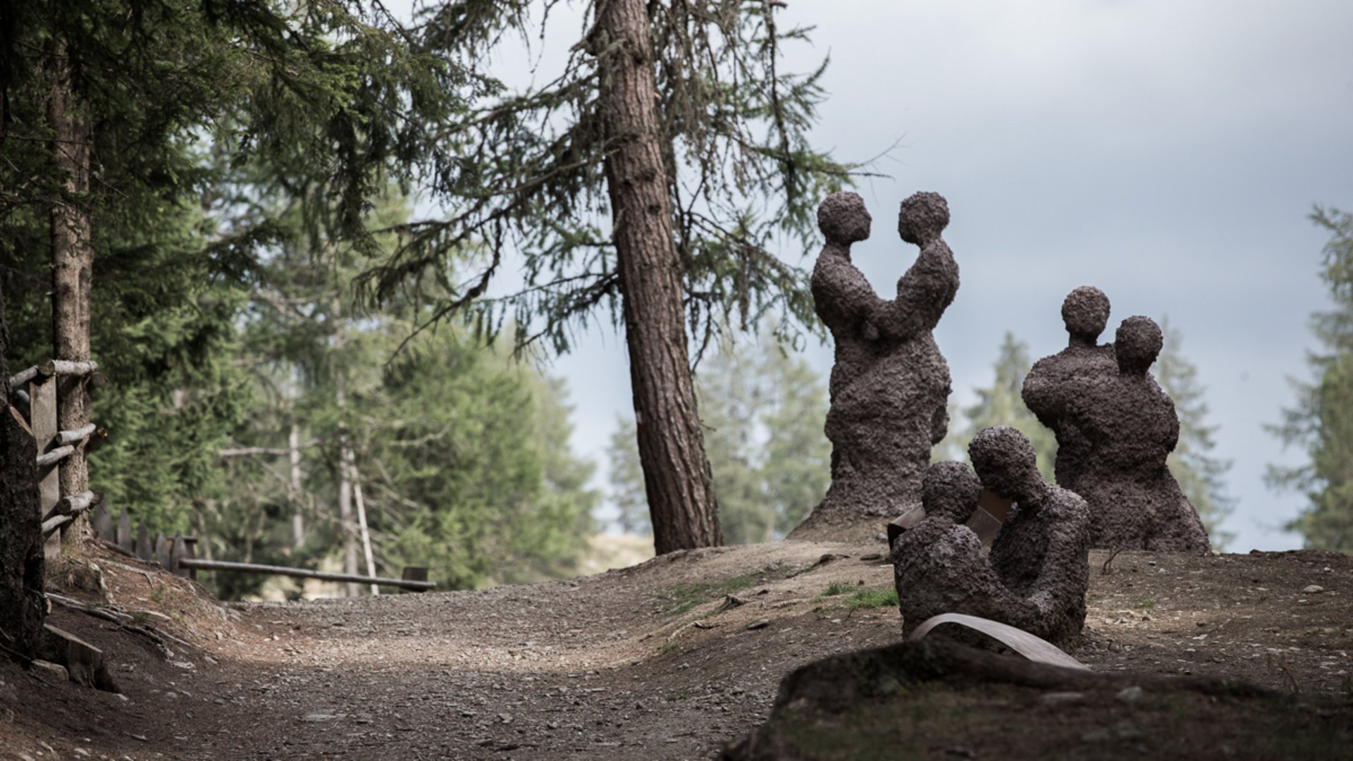 The Creation trail in Rodeneck The picture shows several abstract sculptures of couples, formed from rough material, located on a path in a wooded area. The figures stand or sit close together as if they are embracing or in close contact.