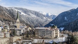 Mühlbach in the Pustertal valley The picture shows a snowy town in a valley, surrounded by forested mountains. In the foreground, there is a church with a tall, pointed tower and several historic buildings, while snow-covered mountains dominate the scene in the background.