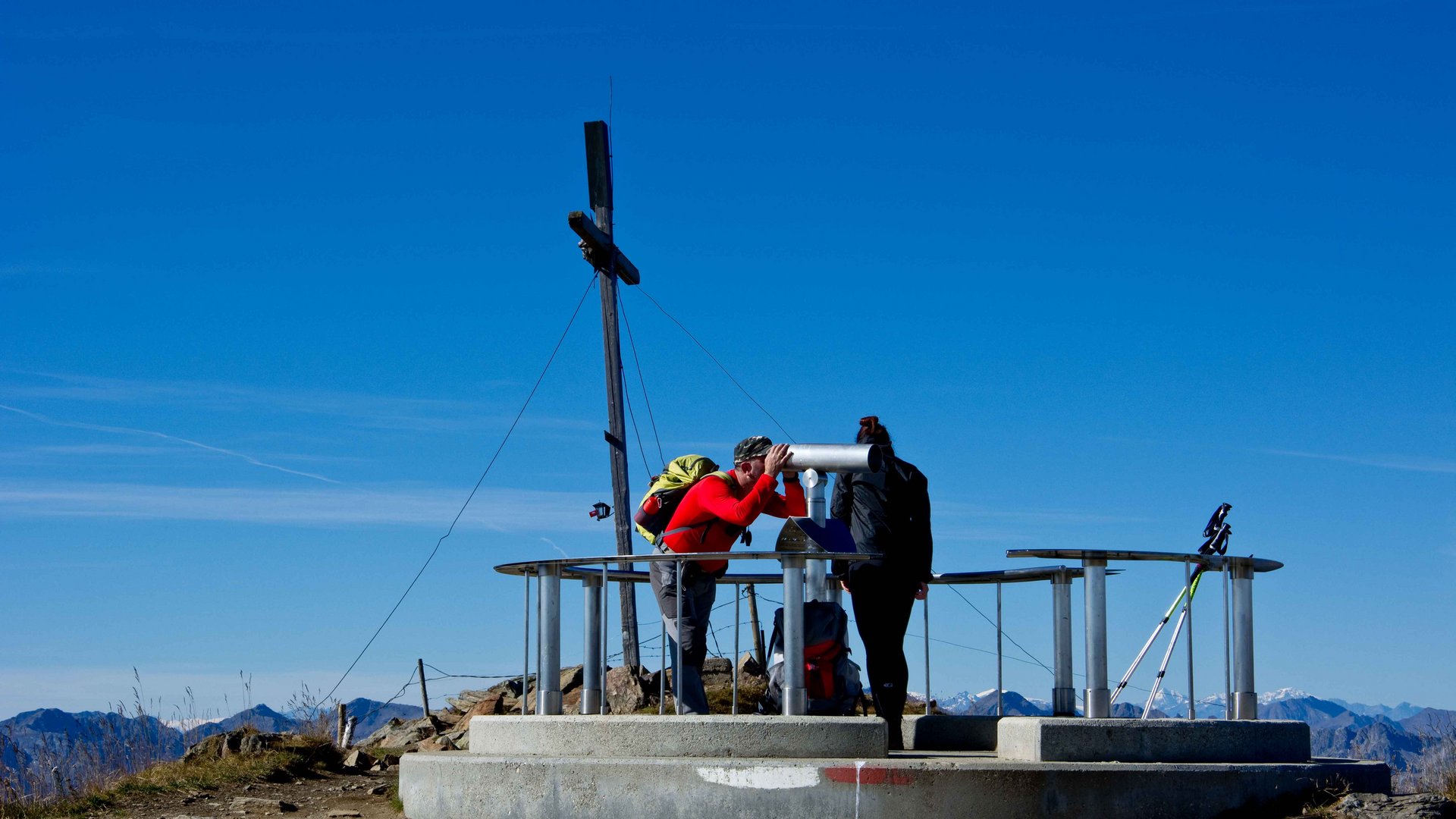 The Gitschberg panoramic platform in Meransen, Italy The picture shows two hikers on a mountain summit looking through a fixed binocular. In the background, there is a large summit cross, and the sky is clear and blue.