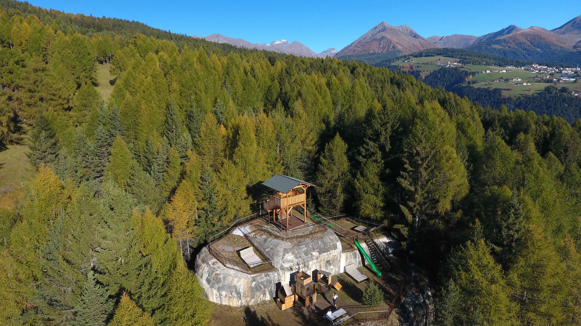 Het recreatiegebied in Spinges/Zuid-Tirol De afbeelding toont een houten uitkijkplatform dat op een rotsrichel ligt te midden van een dicht bos. Op de achtergrond zijn groene bergen en een helderblauwe lucht te zien, die een wijd uitzicht over het landschap bieden.