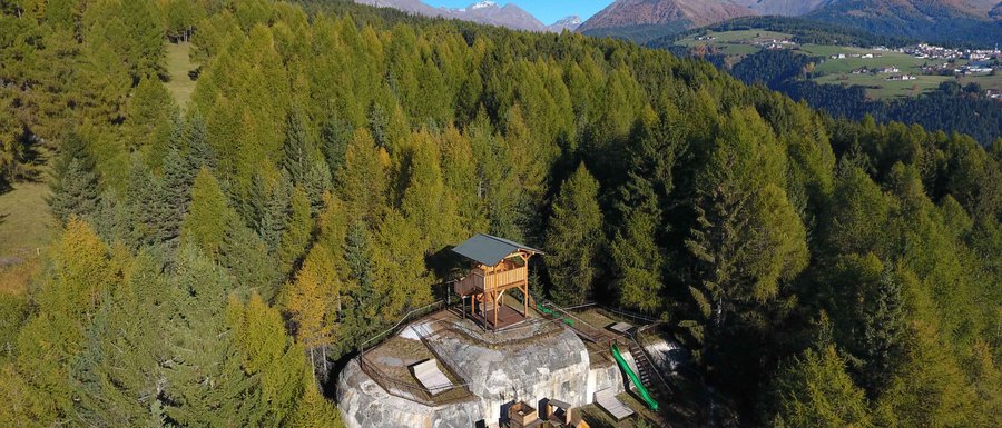 Find your accommodation in South Tyrol The picture shows a wooden observation platform located on a rocky outcrop in the middle of a dense forest. In the background, green mountains and a clear blue sky can be seen, offering a wide view over the landscape.