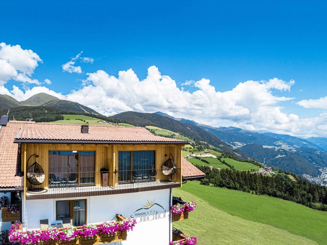 Willkommen in der Ferienregion Gitschberg Jochtal! Das Bild zeigt ein großes Gebäude mit Balkonen, die mit Blumen geschmückt sind, vor einer malerischen Berglandschaft. Im Hintergrund sind grüne Hügel, Wälder und das Tal zu sehen, die sich unter einem strahlend blauen Himmel erstrecken.