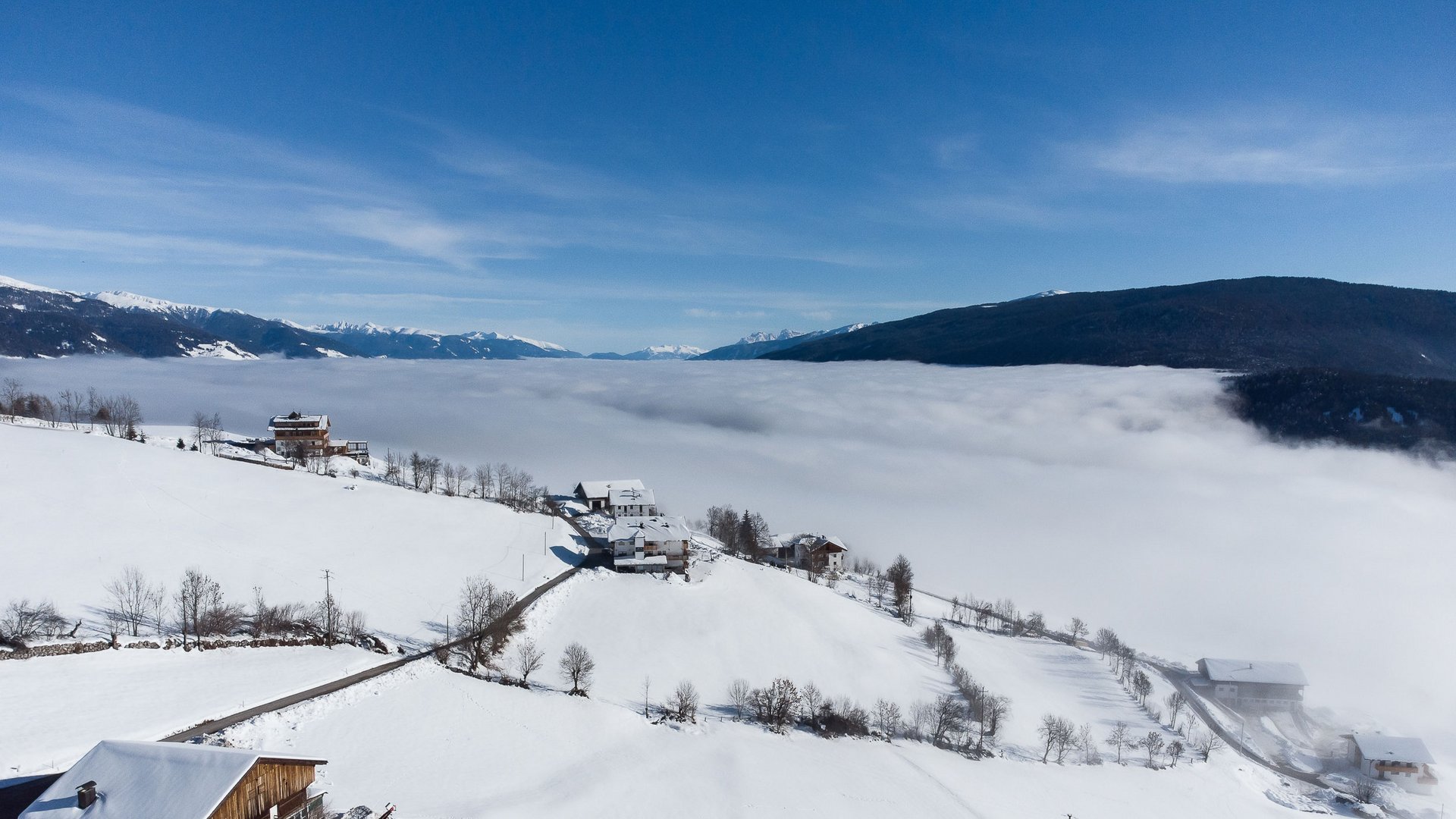 Das Bergdorf Meransen Das Bild zeigt eine winterliche Berglandschaft mit schneebedeckten Feldern und vereinzelten Häusern auf einem Hügel. Im Hintergrund erstreckt sich ein dichtes Wolkenmeer über das Tal, während die schneebedeckten Gipfel der Berge unter einem klaren, blauen Himmel hervorschauen.