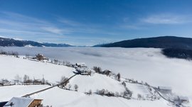 Das Bergdorf Meransen Das Bild zeigt eine winterliche Berglandschaft mit schneebedeckten Feldern und vereinzelten Häusern auf einem Hügel. Im Hintergrund erstreckt sich ein dichtes Wolkenmeer über das Tal, während die schneebedeckten Gipfel der Berge unter einem klaren, blauen Himmel hervorschauen.