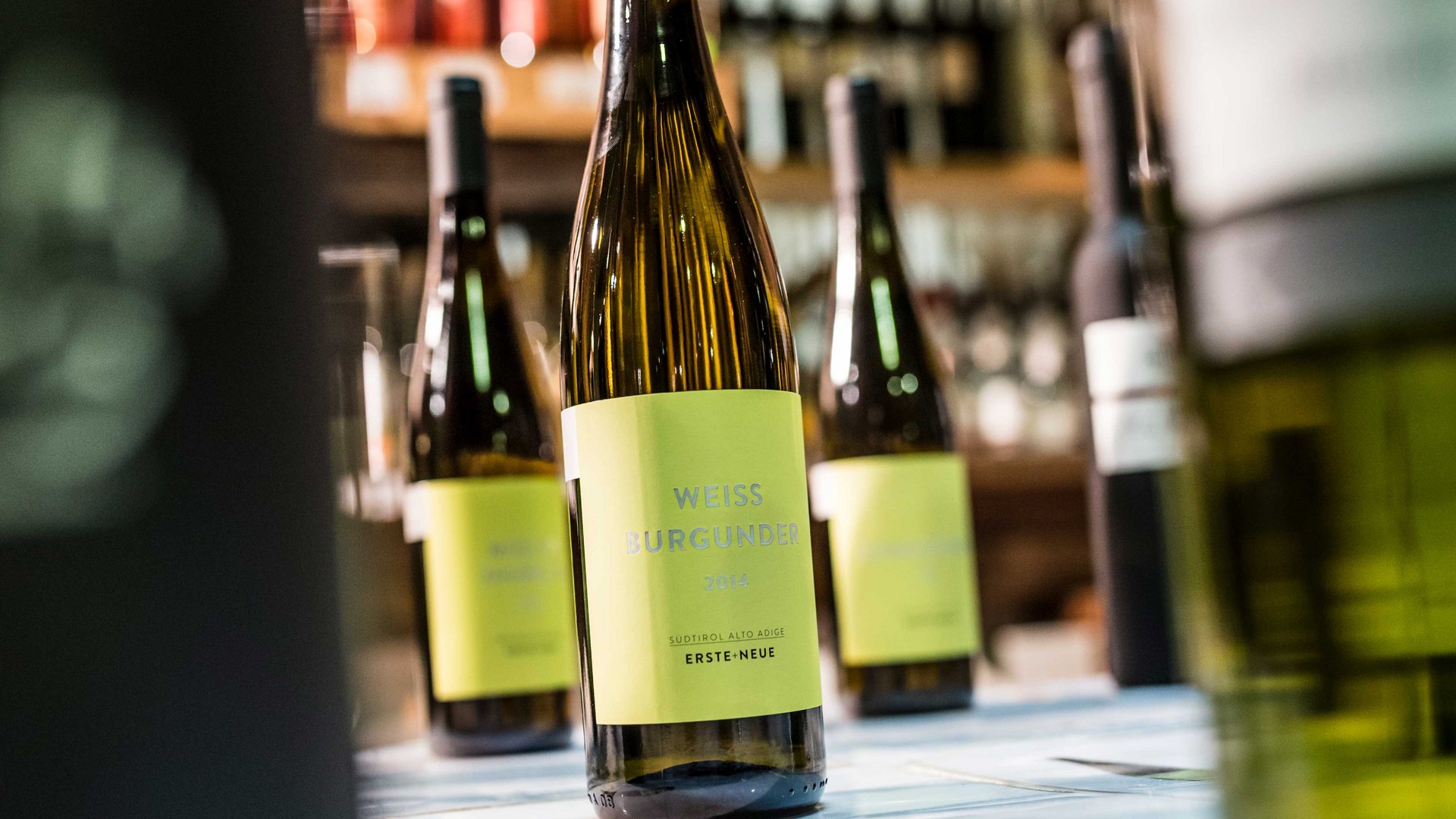 Nakupování v Mühlbachu/Itálie Obrázek ukazuje láhev bílého burgundského vína s žlutou etiketou, na které je napsáno "Weiss Burgunder 2014". V pozadí jsou rozmazaně vidět další lahve vína, které také stojí na polici.