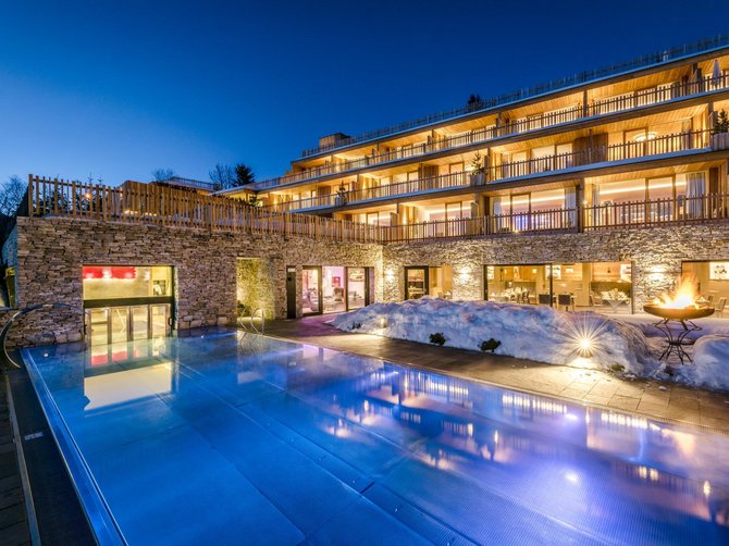 Willkommen in der Ferienregion Gitschberg Jochtal! Das Bild zeigt ein beleuchtetes Hotel in der Dämmerung mit großen Fenstern und Terrassen auf mehreren Etagen. Im Vordergrund befindet sich ein beleuchteter Pool, umgeben von schneebedeckten Flächen und einer Feuerschale, die für eine gemütliche Atmosphäre sorgt.