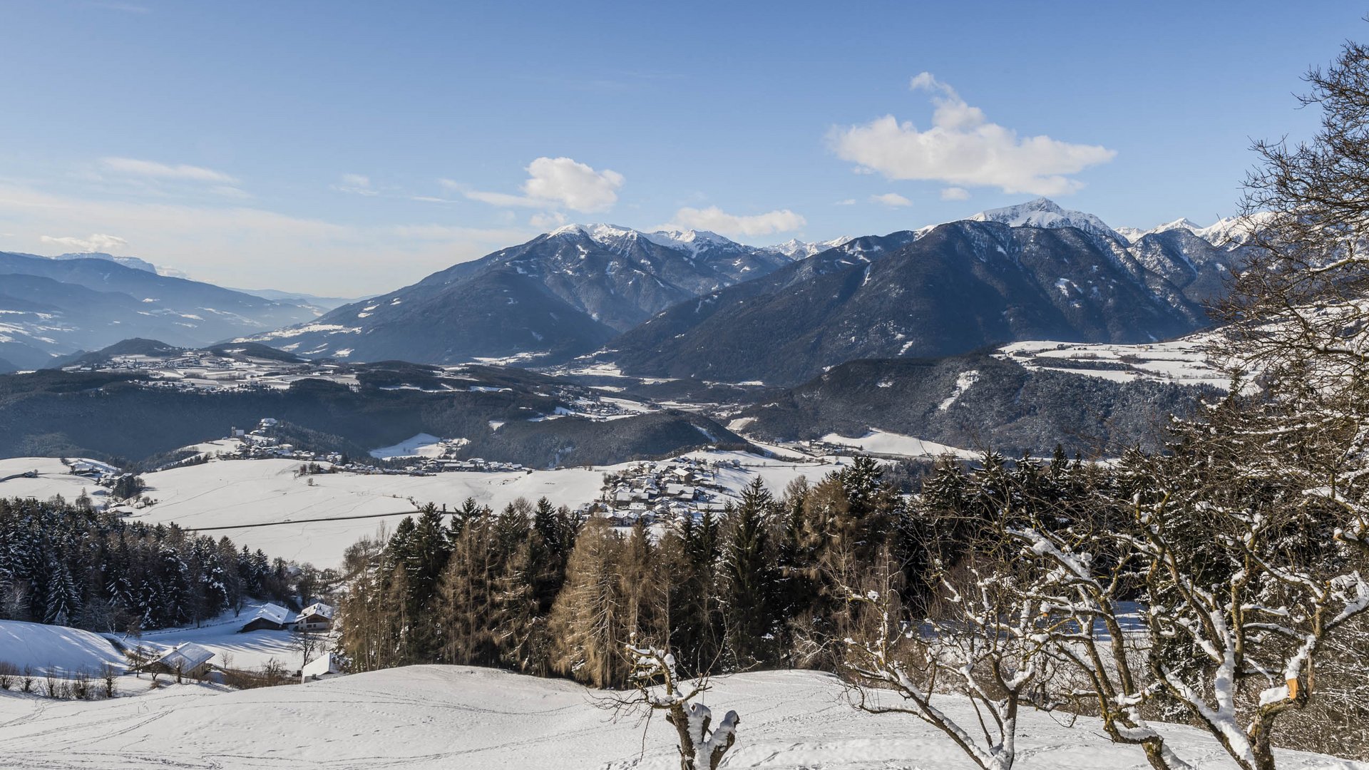 Rodeneck – for a wonderful walking holiday in Italy The picture shows a vast, snow-covered landscape with gentle hills and scattered villages located in a valley. In the background, majestic mountains rise, their peaks also covered with snow, under a clear blue sky.