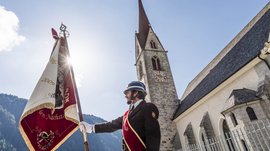 Vals: das Almendorf Das Bild zeigt einen Mann in einer traditionellen Uniform mit Helm, der eine große Fahne hält. Im Hintergrund ist eine Kirche mit einem hohen Turm und Uhr zu sehen, während die Sonne hinter der Fahne hervorscheint.