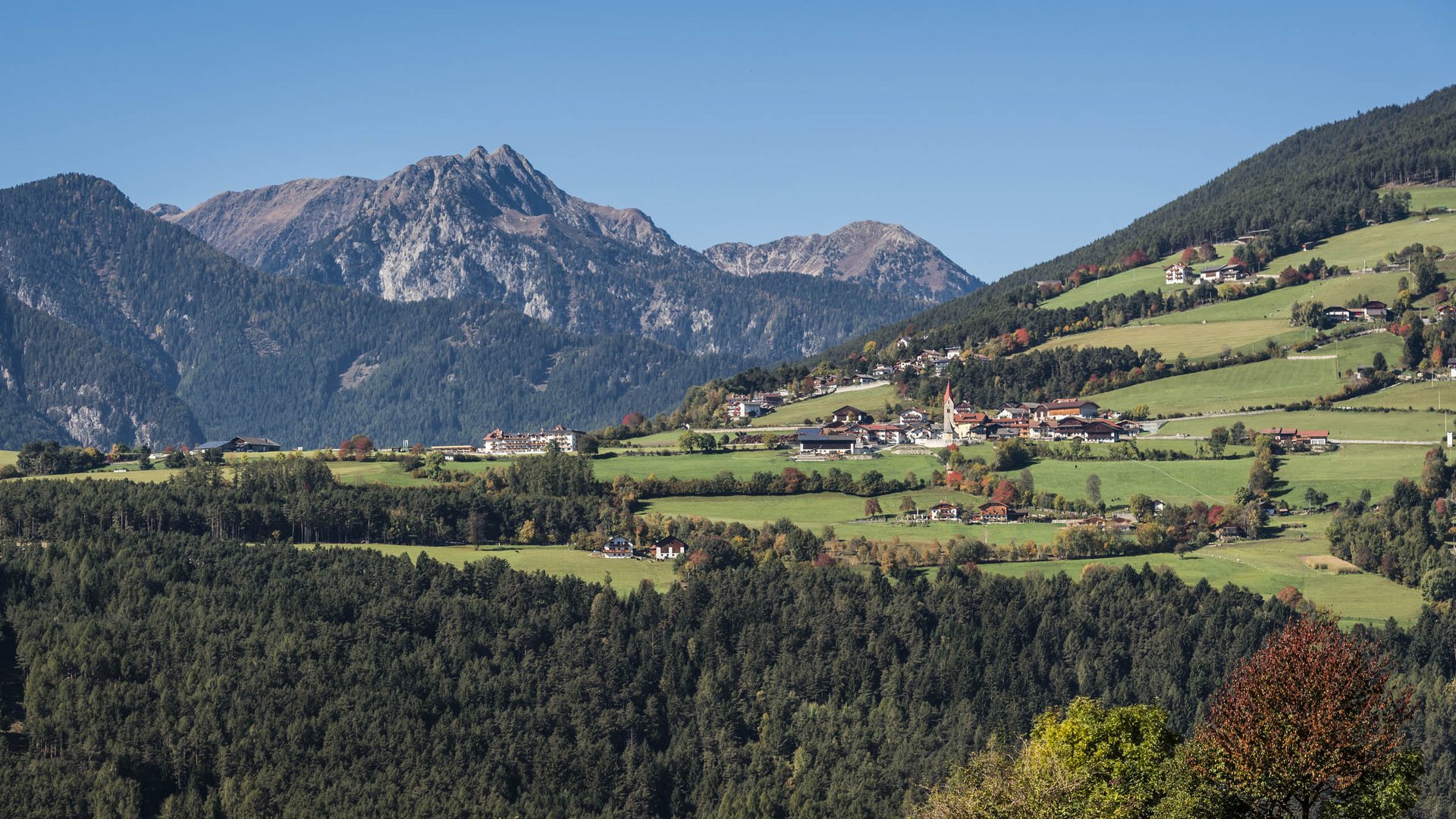 The Alpine hamlet of Spinges The picture shows a green, hilly landscape with scattered houses and a church tower in the middle. In the background, impressive mountains rise up, framing the scene.