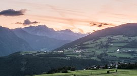 The Alpine hamlet of Spinges The picture shows a gentle hilly landscape at sunset, with a rural area in the foreground. In the background, dark mountain ranges are visible, glowing in pink and orange hues in the soft light of the evening sky.