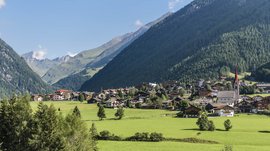 Vals: das Almendorf Das Bild zeigt ein malerisches Bergdorf mit Häusern, die sich in ein grünes Tal schmiegen. Im Hintergrund ragen bewaldete Berghänge auf, während eine Kirche mit rotem Turm die Landschaft prägt.