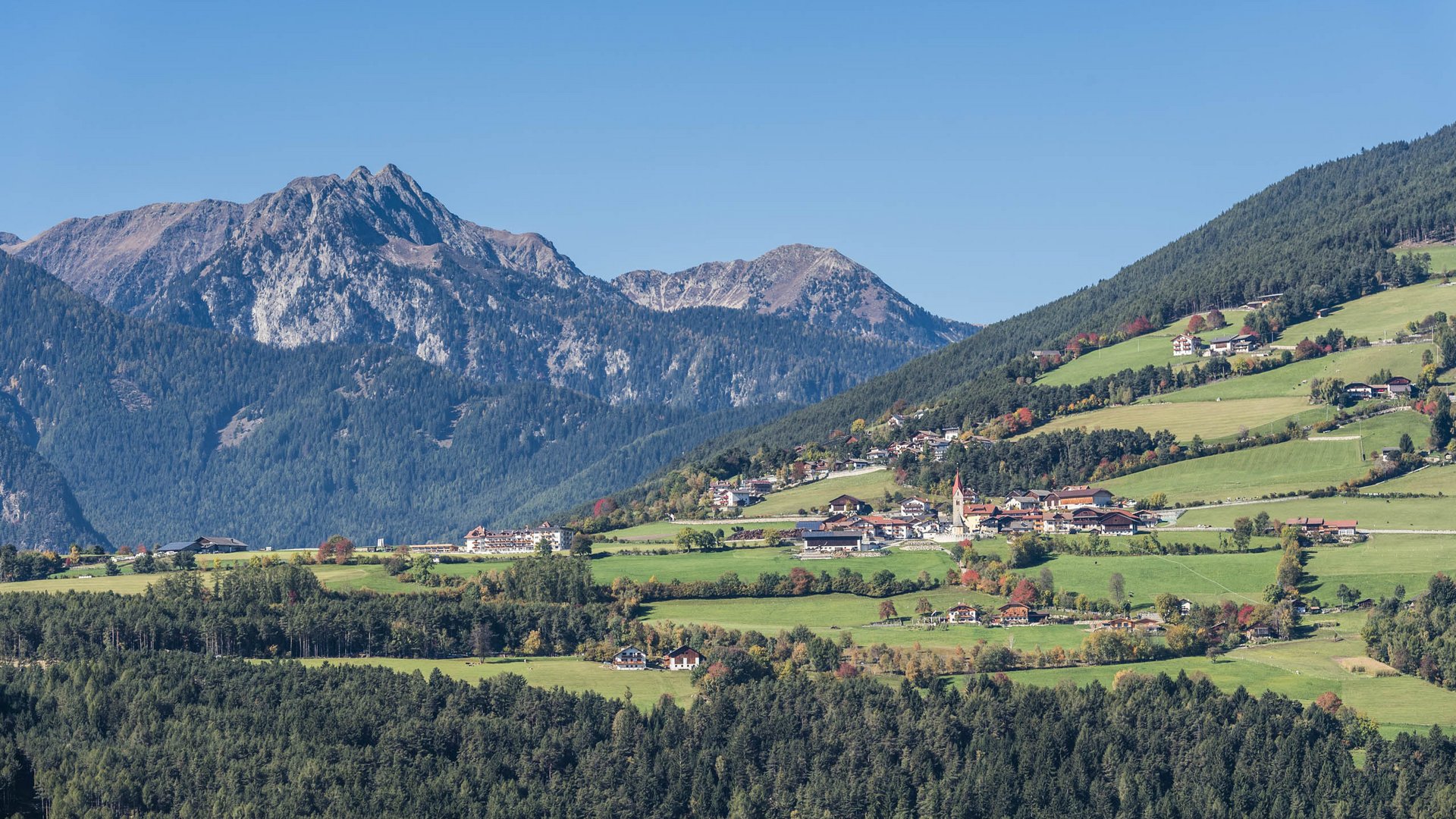 The Alpine hamlet of Spinges The picture shows an idyllic landscape with green hills, scattered with small houses and a church with a red tower. In the background, impressive mountains rise, covered by dense forests.