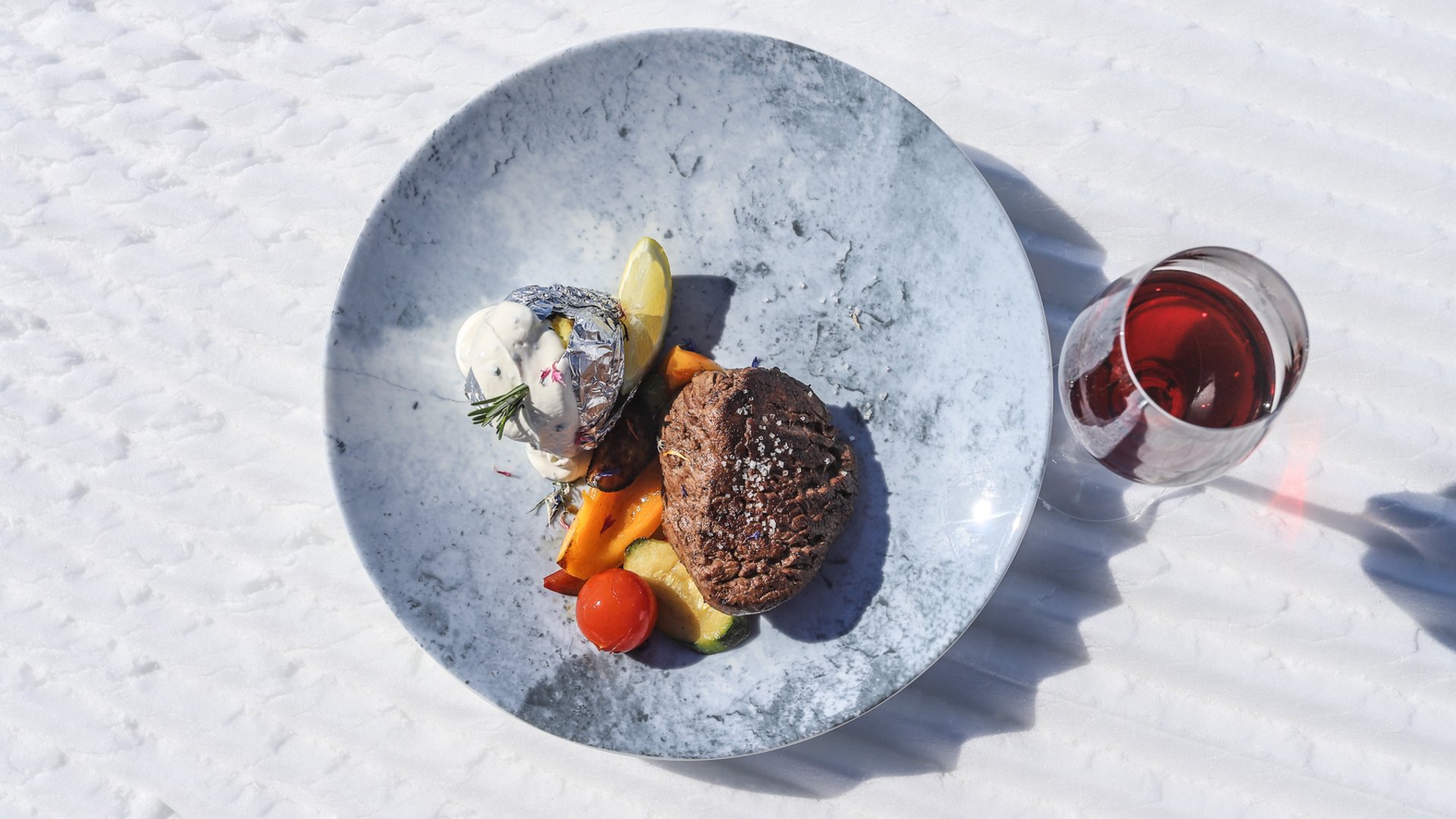 Gitschberg Jochtal holiday area – mountain huts and shelters In the picture, there is an appetizingly arranged plate with a grilled steak, accompanied by grilled vegetables and a baked potato wrapped in aluminum foil with herb cream, served on a marbled plate on a snow-covered surface next to a glass of red wine.