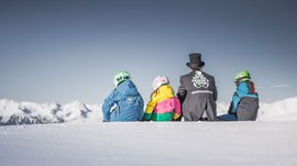 Skigebied Gitschberg Jochtal Drie kinderen en een volwassene zitten in de sneeuw met uitzicht op bergen