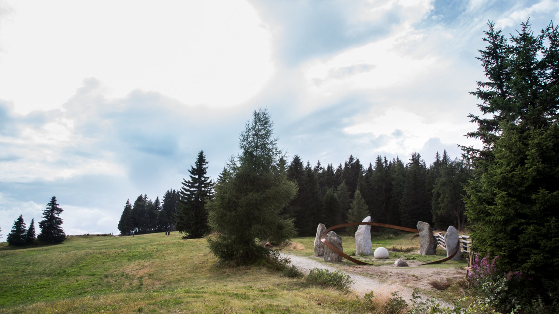 The Creation trail in Rodeneck The image shows a grassy landscape with scattered trees in the foreground and a dense forest in the background. In a clearing, there is an installation made up of several large stones surrounded by a metallic ring.