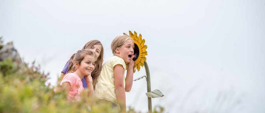 Find your accommodation in South Tyrol The picture shows three smiling children leaning against a large, yellow sunflower. The sunflower appears to be part of an installation or playground, and the children seem to be having fun interacting with it.