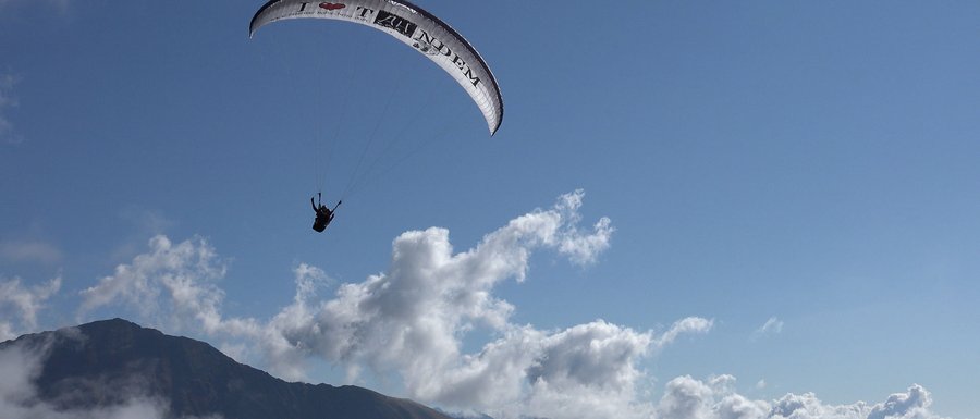 Find your accommodation in South Tyrol The picture shows a paraglider soaring high above a mountain landscape. The sky is clear and blue, with a few scattered clouds, and you can see the silhouette of a mountain in the background.