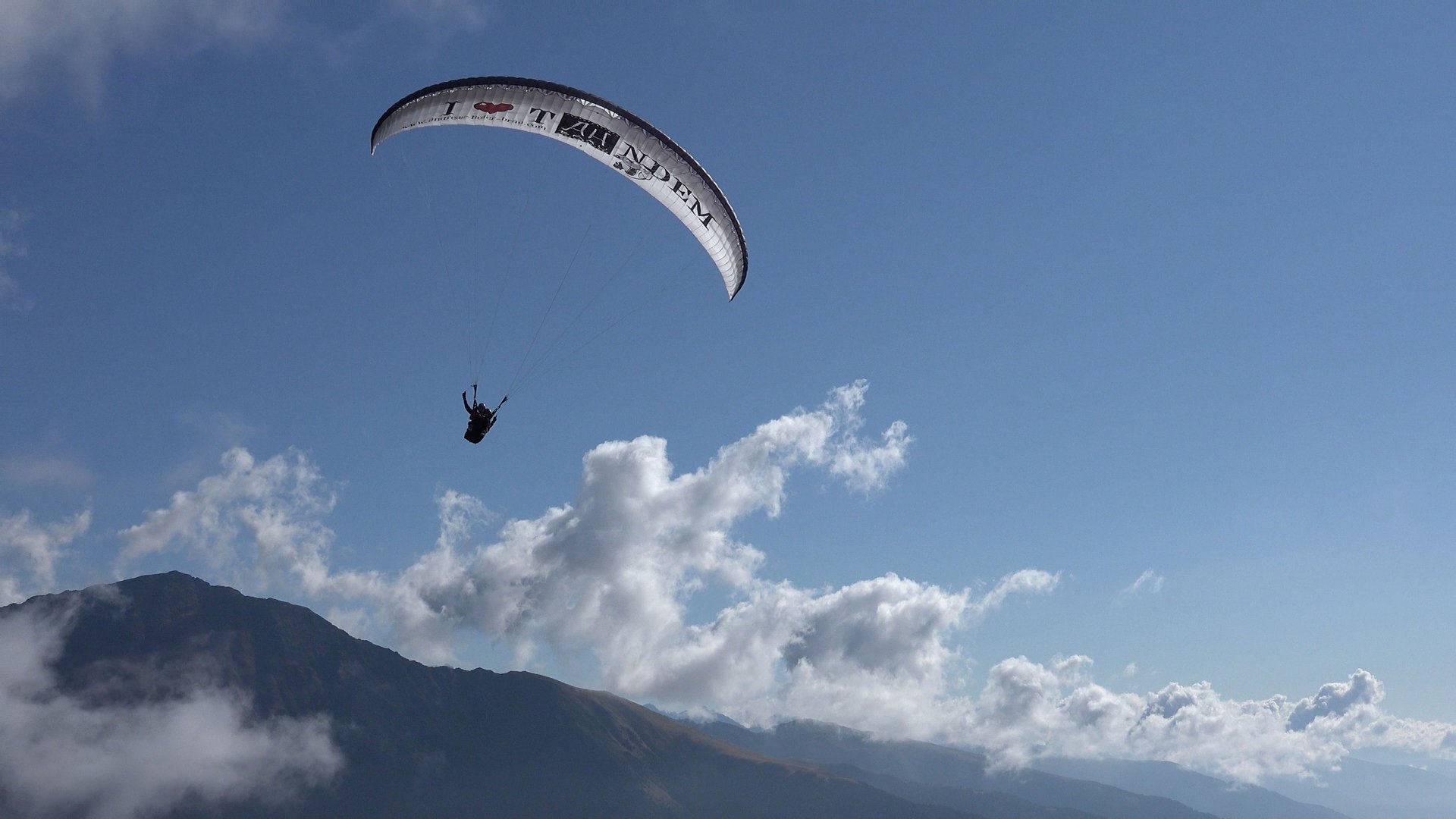 Paragliden in Zuid-Tirol De afbeelding toont een paraglider die hoog boven een berglandschap zweeft. De lucht is helder en blauw, met een paar verspreide wolken, en je kunt de silhouet van een berg op de achtergrond zien.