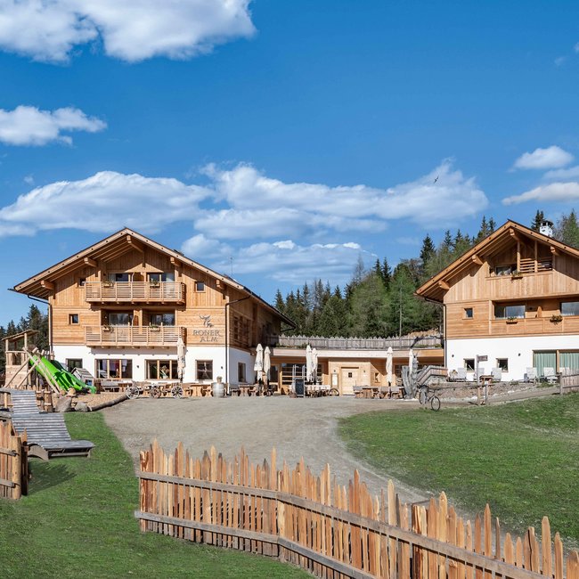 Naturhotel Runa Obrázek ukazuje dvě moderní dřevěné budovy "Roner Alm" pod jasnou modrou oblohou s ojedinělými bílými mraky. Budovy jsou obklopeny dřevěným plotem a v popředí se nachází dětské hřiště se skluzavkou a několika dřevěnými lavičkami. V pozadí jsou vidět lesy a kopce.