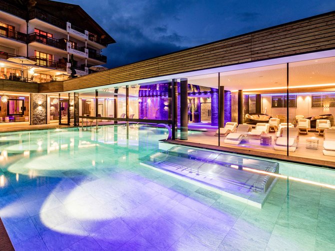 Willkommen in der Ferienregion Gitschberg Jochtal! Das Bild zeigt einen beleuchteten Außenpool bei Nacht, umgeben von einem modernen Gebäude mit großen Glaswänden. Der Pool leuchtet in verschiedenen Farben, während innen weiße Liegen und eine gemütliche Atmosphäre zu sehen sind.