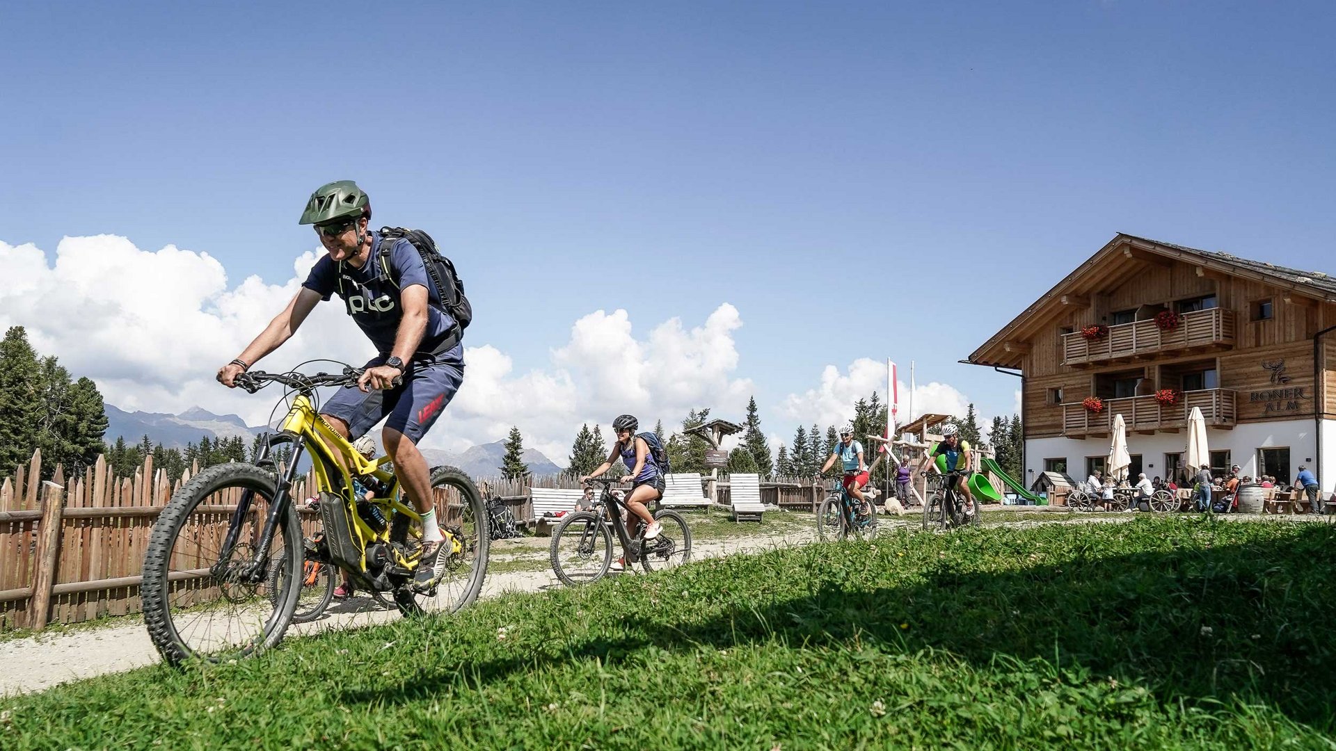 De Rodenecker-Lüsner Alm De afbeelding toont een groep mensen die met mountainbikes een pad volgen. Op de achtergrond is een houten gebouw te zien dat lijkt op een berghut, en er zijn meer mensen die daar verblijven. De lucht is helder en blauw, en het weer lijkt zonnig te zijn.