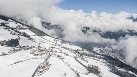 The Alpine hamlet of Spinges The picture shows a snow-covered village in the mountains, nestled in hilly landscapes. Clouds envelop the surrounding valleys and peaks, while a clear blue sky is visible above the village.
