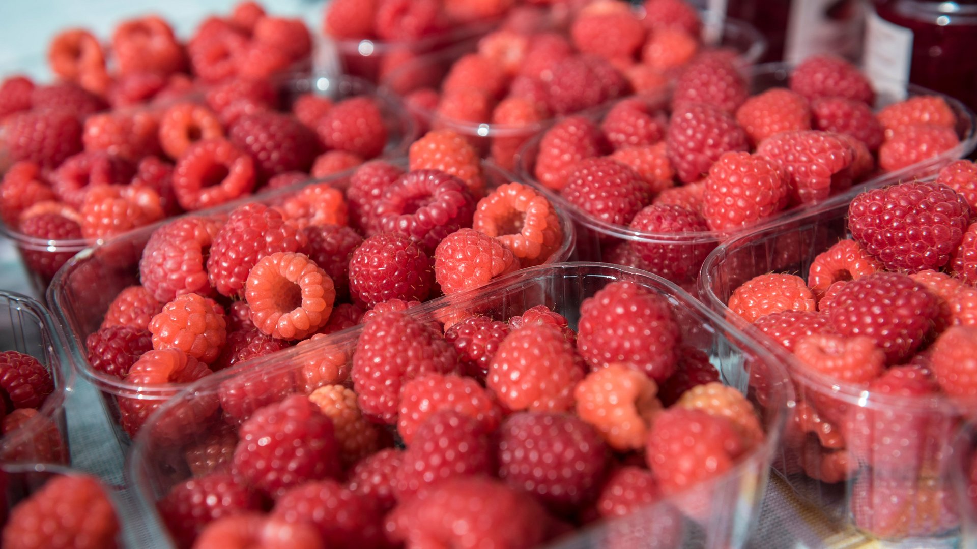 Nakupování v Mühlbachu/Itálie Obrázek ukazuje několik průhledných plastových misek naplněných čerstvými červenými malinami. Maliny jsou na obrázku vidět zblízka a jasně, což zdůrazňuje jejich jasnou barvu a šťavnatou texturu.