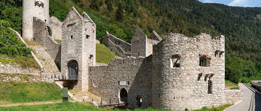 Find your accommodation in South Tyrol The picture shows an old stone castle ruin with several well-preserved towers and walls. The castle is located on a mountainside, surrounded by green meadows and dense forests, under a clear blue sky.