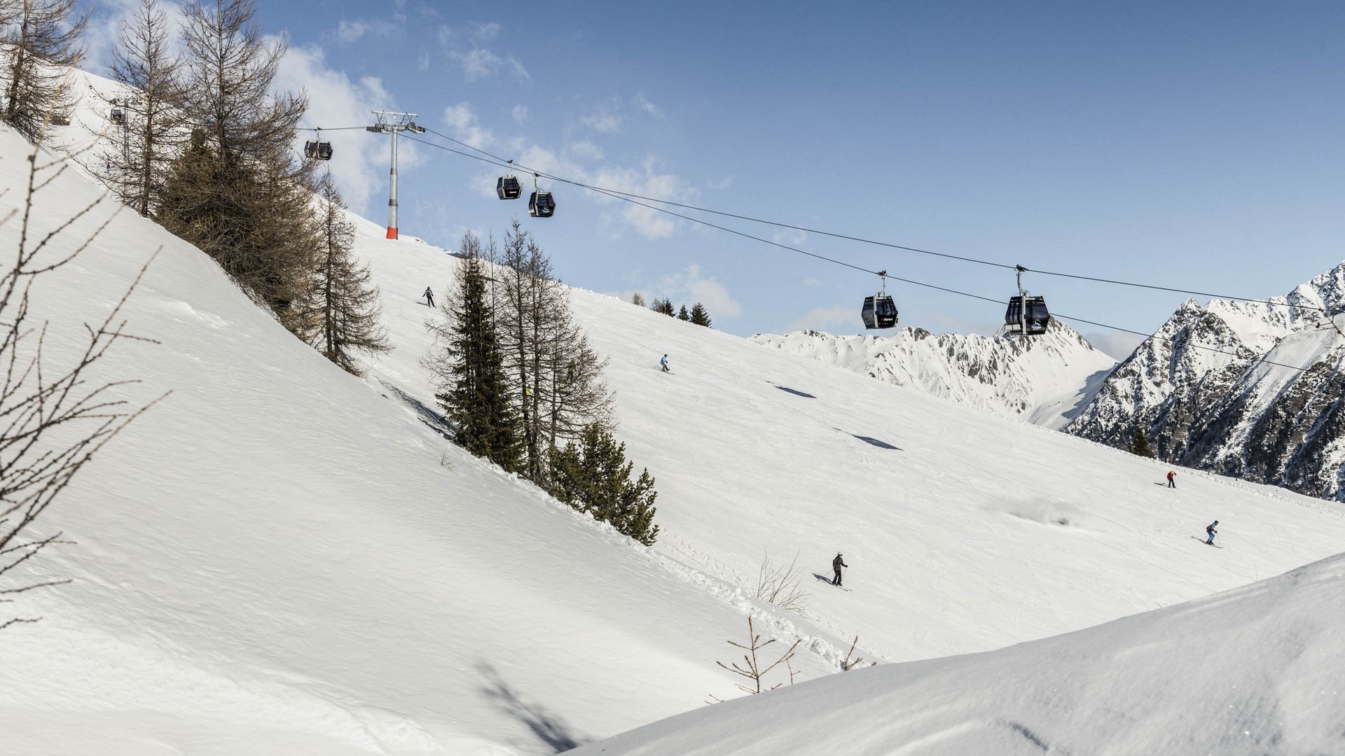 Skigebied Gitschberg Jochtal De afbeelding toont een besneeuwde skipiste met meerdere skiërs die de helling afrijden. Daarboven loopt een kabelbaan met gondels die skiërs de berg op vervoert. Op de achtergrond zijn besneeuwde bergen en een heldere, blauwe lucht te zien.