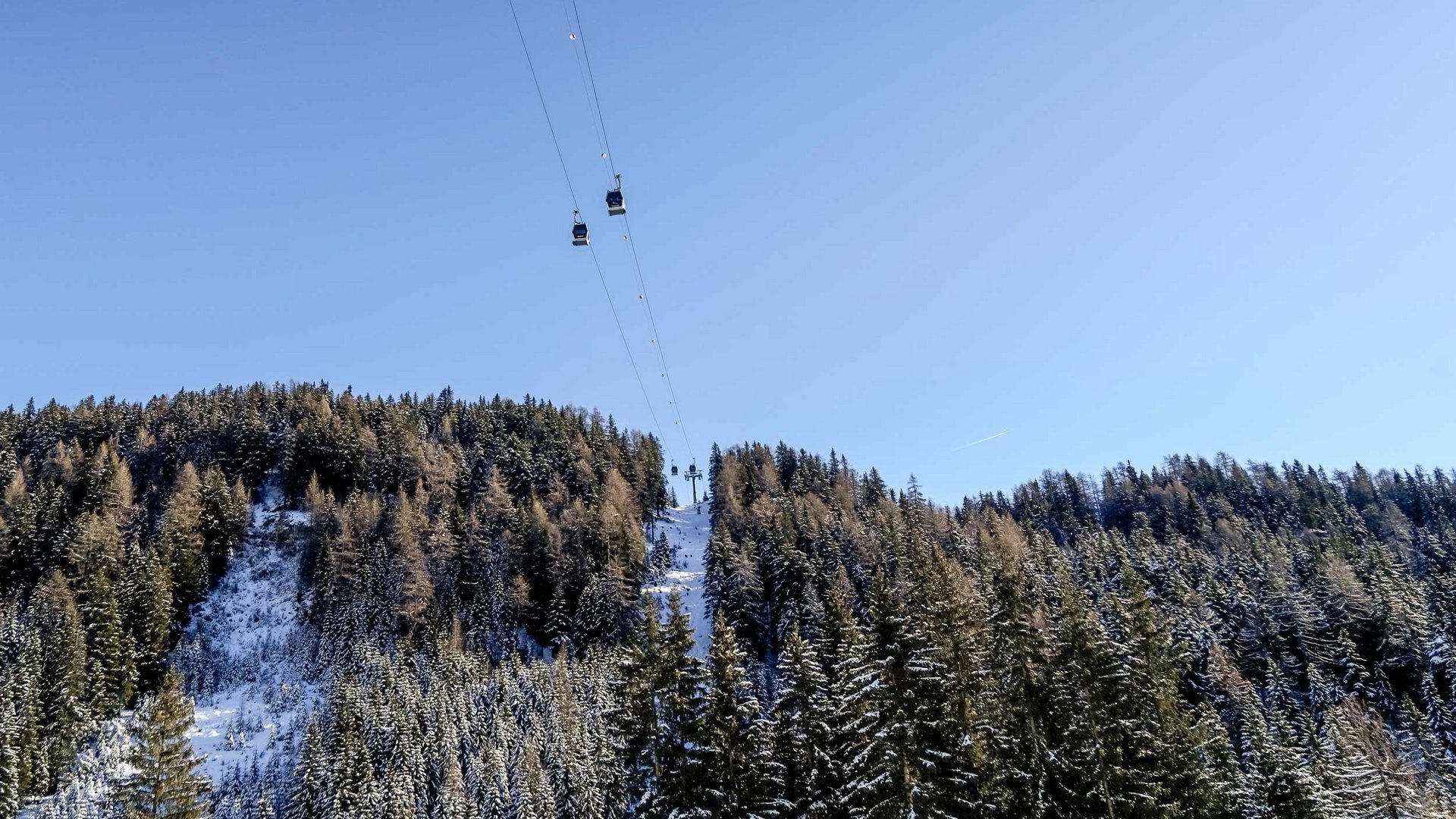 Het Altfassdal bij Meransen De afbeelding toont een kabelbaan met gondels die over met sneeuw bedekte bomen en een beboste heuvel gaat. De lucht is helder en blauw, en de kabelbaan leidt naar een top die ook met sneeuw bedekt is.