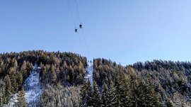 Het Altfassdal bij Meransen De afbeelding toont een kabelbaan met gondels die over met sneeuw bedekte bomen en een beboste heuvel gaat. De lucht is helder en blauw, en de kabelbaan leidt naar een top die ook met sneeuw bedekt is.