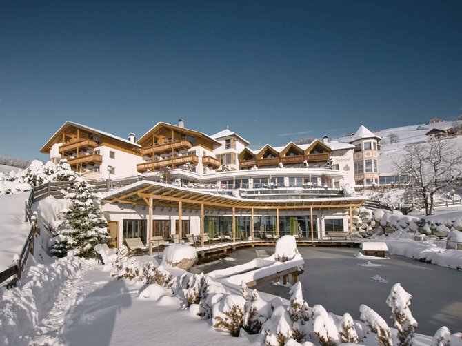 Willkommen in der Ferienregion Gitschberg Jochtal! Das Bild zeigt ein großes Hotel in einer verschneiten Winterlandschaft. Vor dem Hotel liegt ein zugefrorener Teich, und die Dächer sowie die Umgebung sind dick mit Schnee bedeckt.