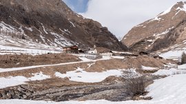 Fane Alm – nejkrásnější horská vesnice Jižního Tyrolska Obrázek zobrazuje zimní horskou krajinu s malou vesnicí obklopenou zasněženými a částečně zasněženými svahy. V popředí teče potok a v pozadí je vidět několik dřevěných chat a malý kostel.